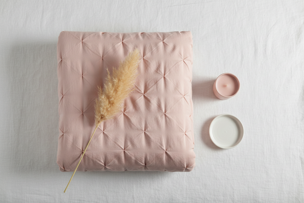 Flat lay of folded blush pintuck duvet cover with pampas grass, pink candle and ceramic dish