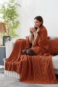 Woman wrapped in rust cable knit throw on a sofa with matching cushion covers and a warm drink